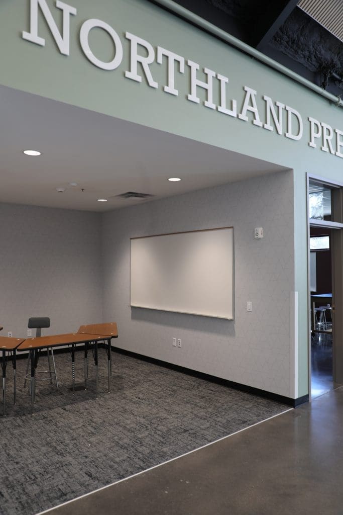 A modern classroom corner in the Northland Preparatory Academy STEAM Building features a blank whiteboard, a wooden table, a chair, and part of a sign reading "NORTHLAND PRE" on the wall above.