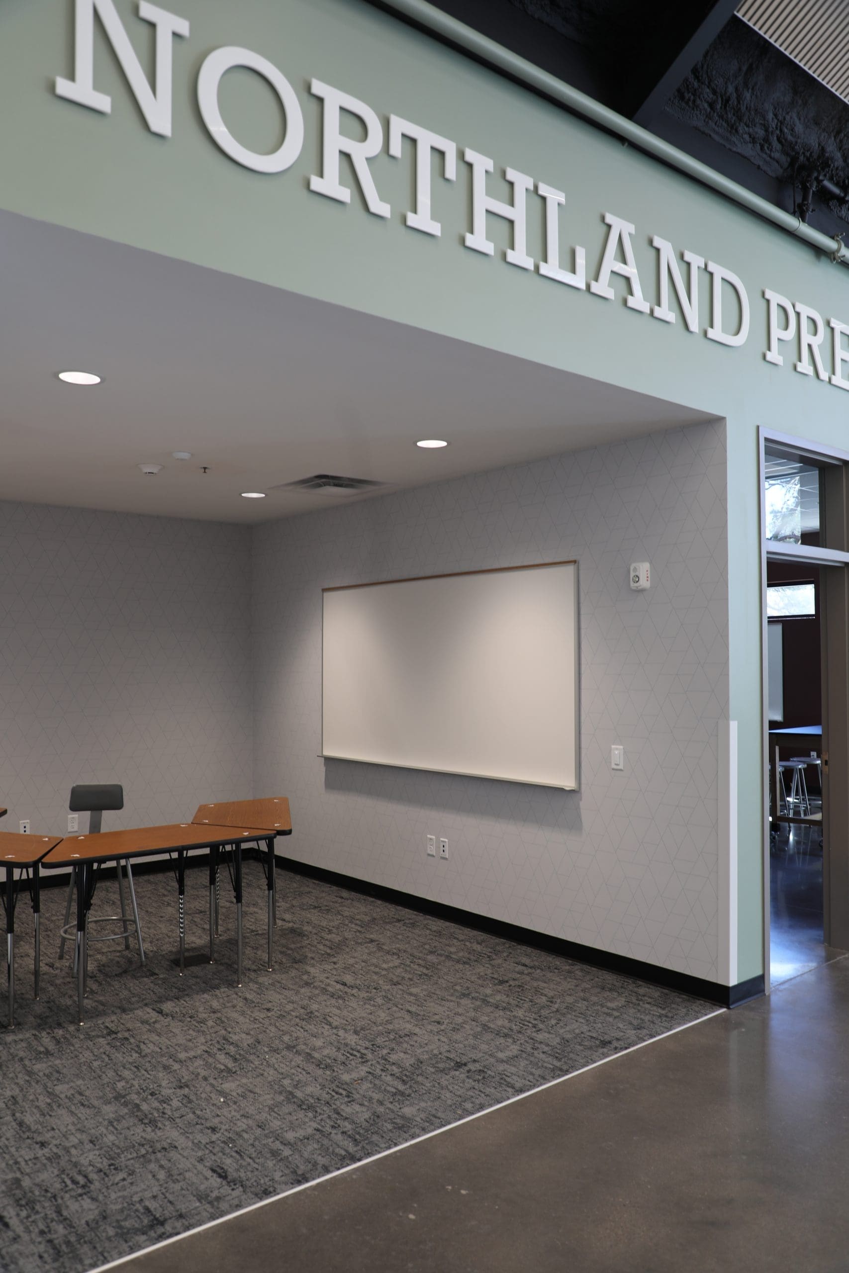 A modern classroom corner in the Northland Preparatory Academy STEAM Building features a blank whiteboard, a wooden table, a chair, and part of a sign reading "NORTHLAND PRE" on the wall above.