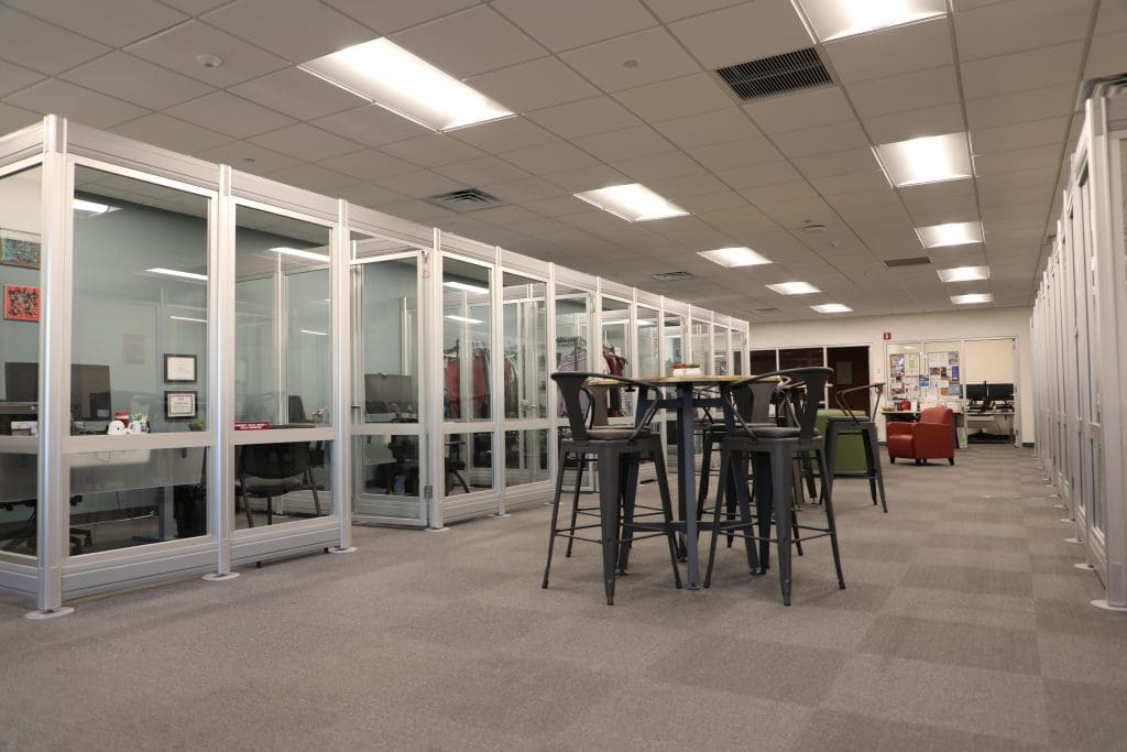 Office space with glass-walled cubicles along one side, high tables with tall chairs in the center, and lounge seating in the background.