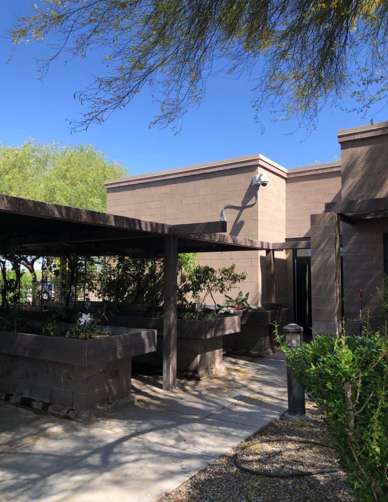 A modern brown brick building with a covered walkway, surrounded by greenery and trees under a clear blue sky.
