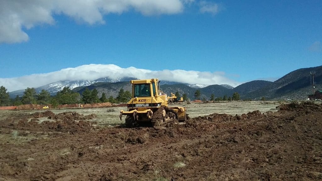 A yellow bulldozer moves earth on a muddy field with mountains and trees in the background under a partly cloudy sky.