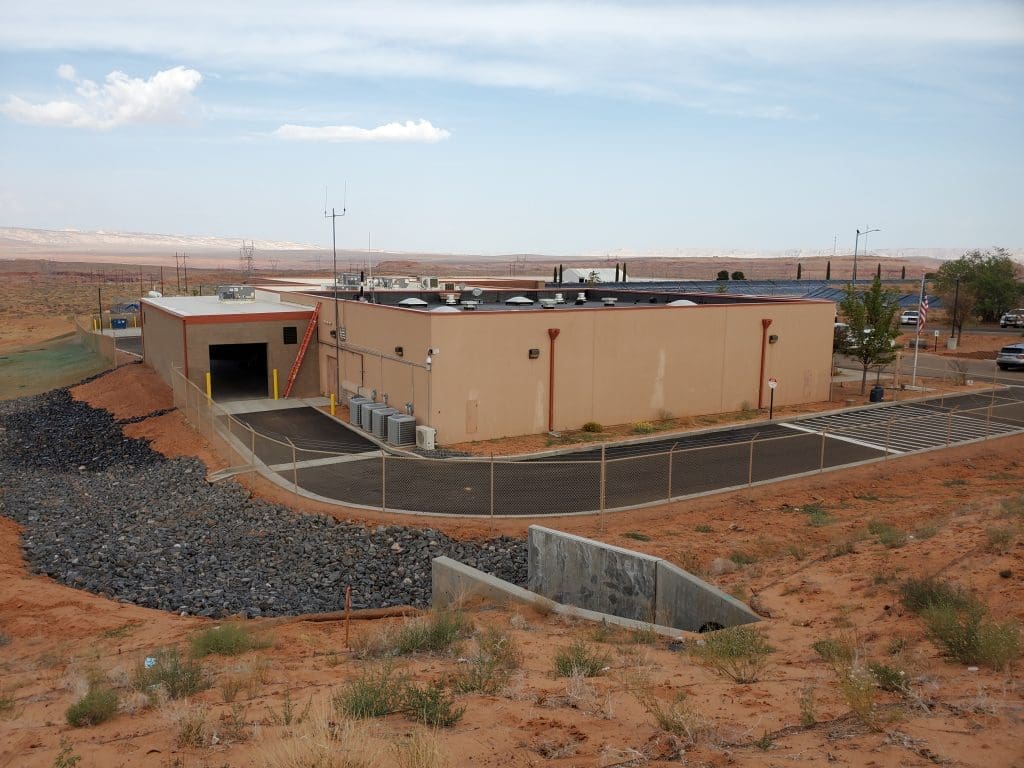 A low, rectangular beige building with rooftop equipment is surrounded by a fence, set in a dry, desert landscape with rocky ground and sparse vegetation.