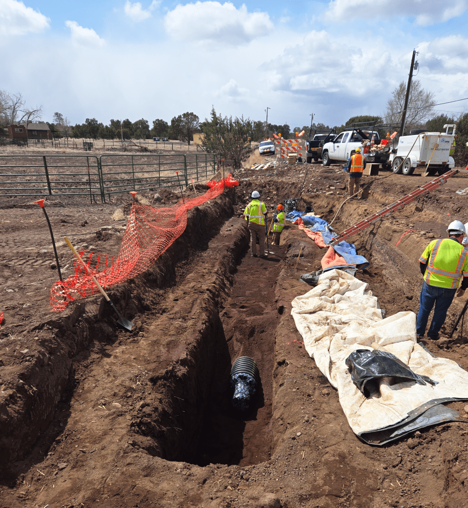 Construction workers in safety gear work in and around a deep trench with a large pipe, surrounded by equipment, vehicles, and orange safety fencing on a dirt site.