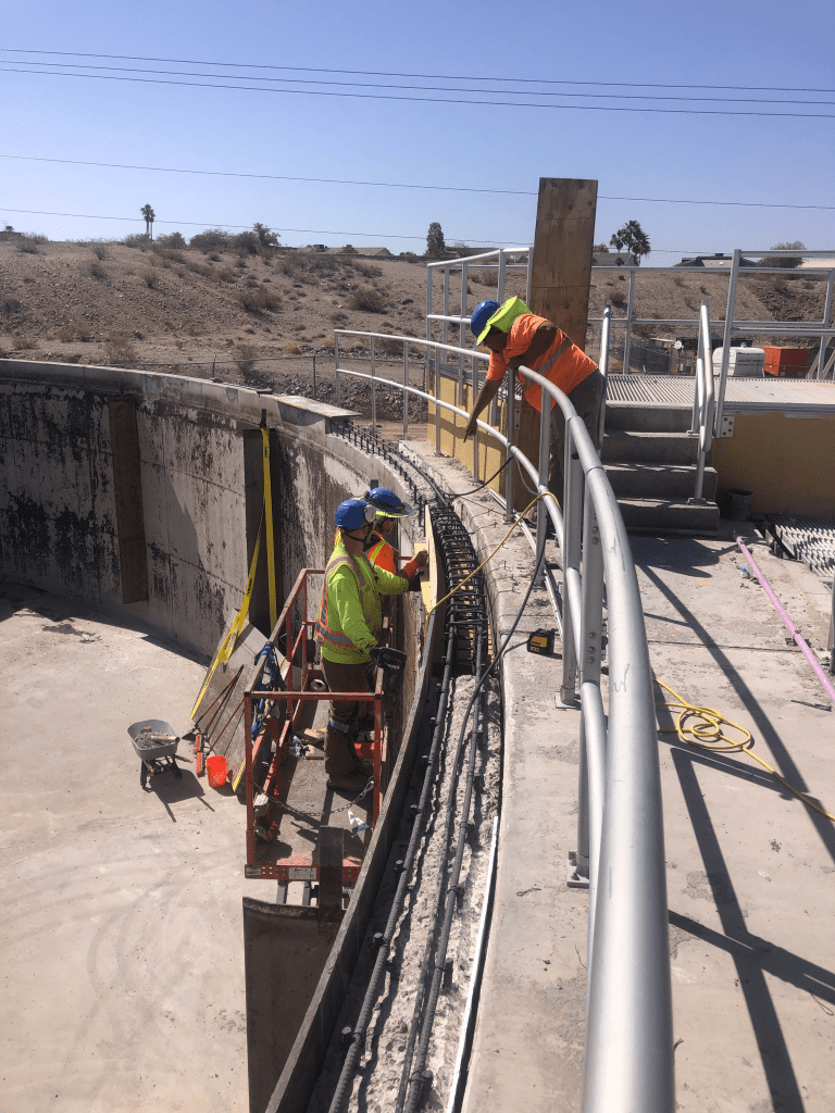 Two workers on a lift and one on stairs perform maintenance or repairs on the curved edge of a large outdoor concrete structure under clear skies.