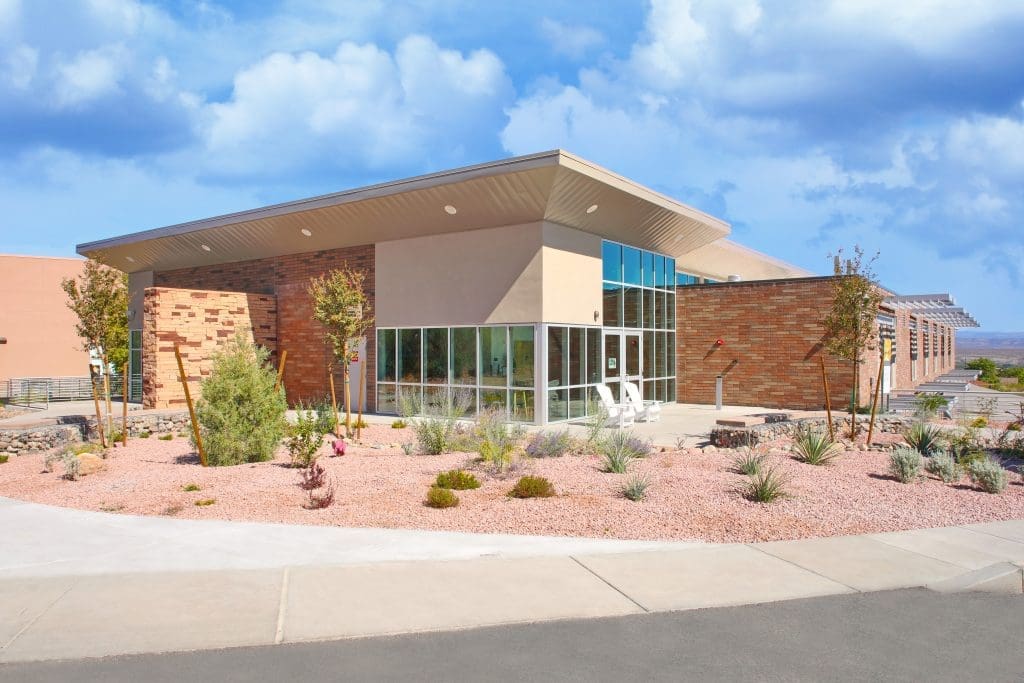 Modern, single-story building with large windows and stone accents, surrounded by desert landscaping under a partly cloudy sky.