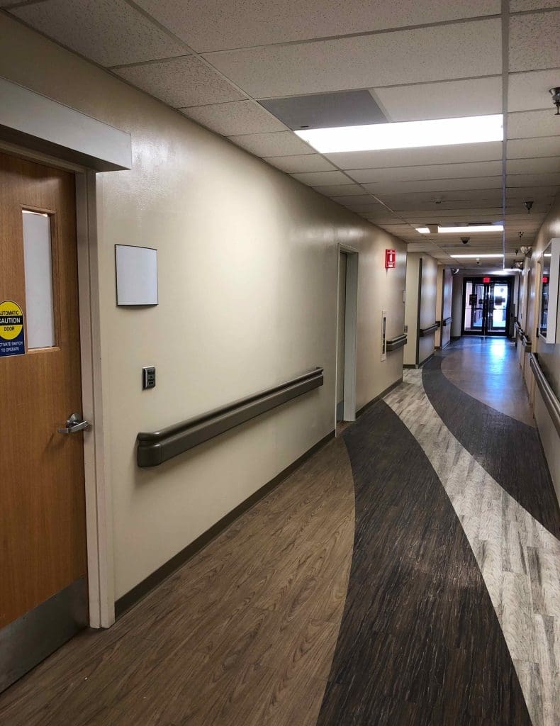 Hospital hallway with beige walls, wood-style flooring, handrails, and a closed door on the left; corridor leads to glass doors at the far end.