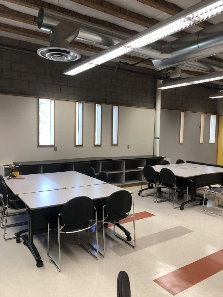 A classroom with several tables and black chairs, white walls with narrow windows, exposed ceiling pipes, and built-in shelves along one wall.