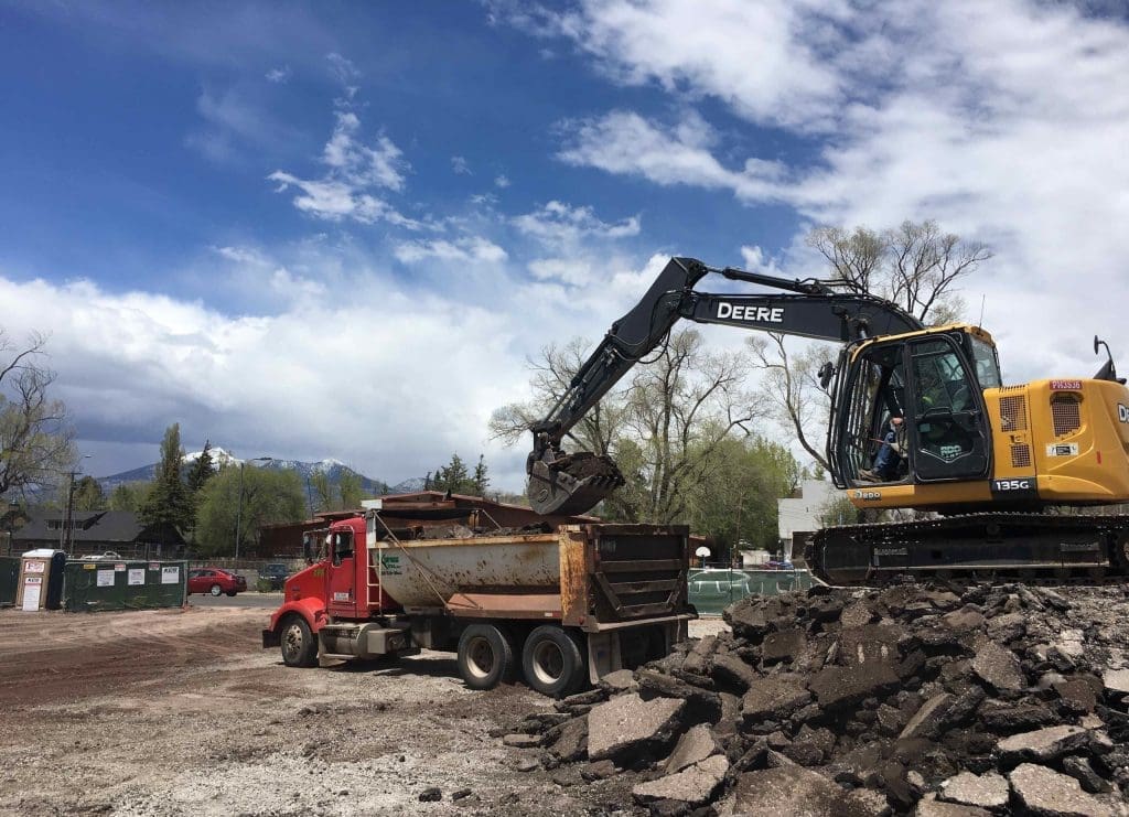An excavator loads rubble into a red dump truck at a construction site under a partly cloudy sky.