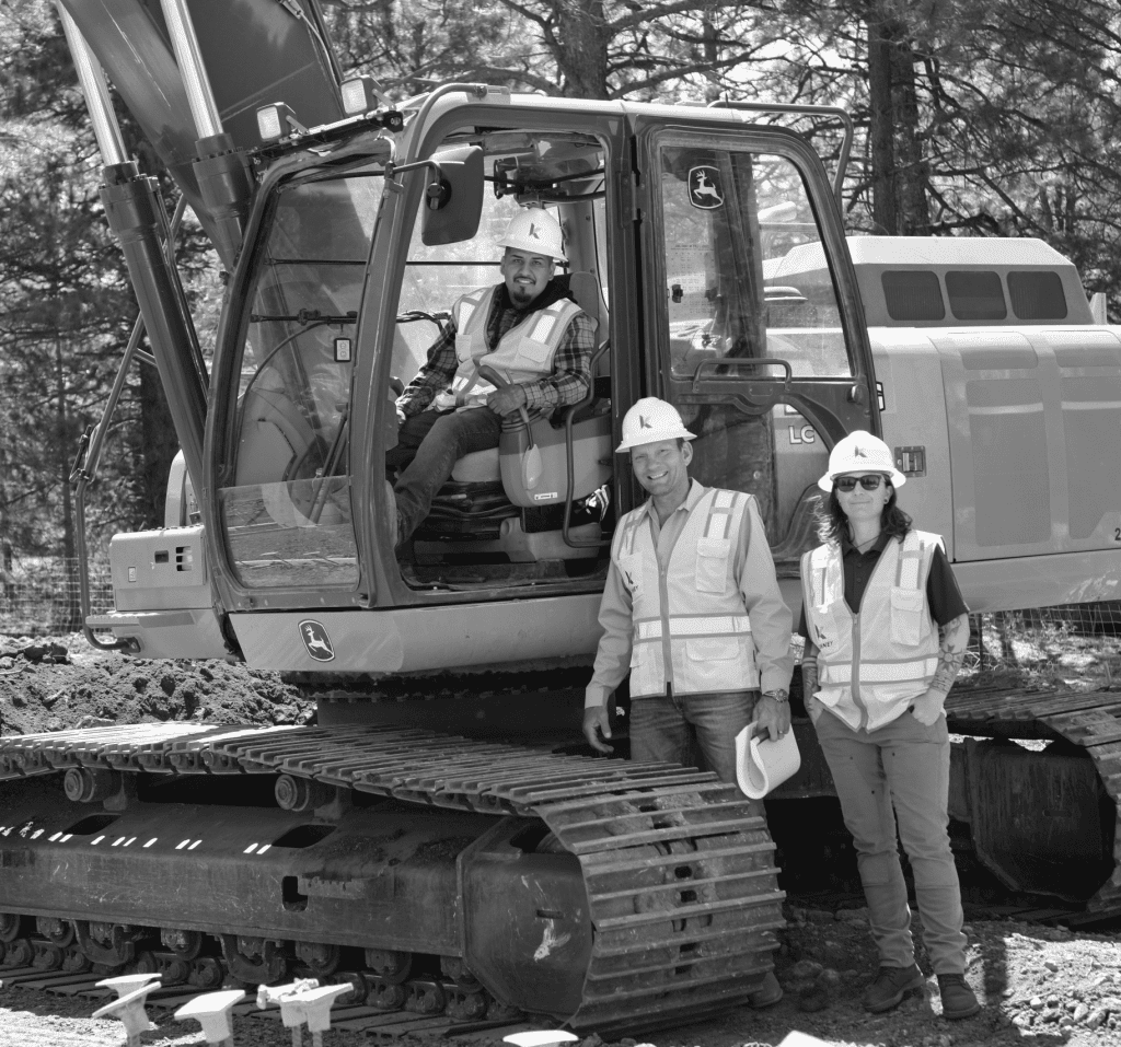 Three people wearing safety vests and hard hats are at a construction site, with one sitting inside an excavator and two standing in front of it, posing for the photo—showcasing a day on the job with About Kinney.