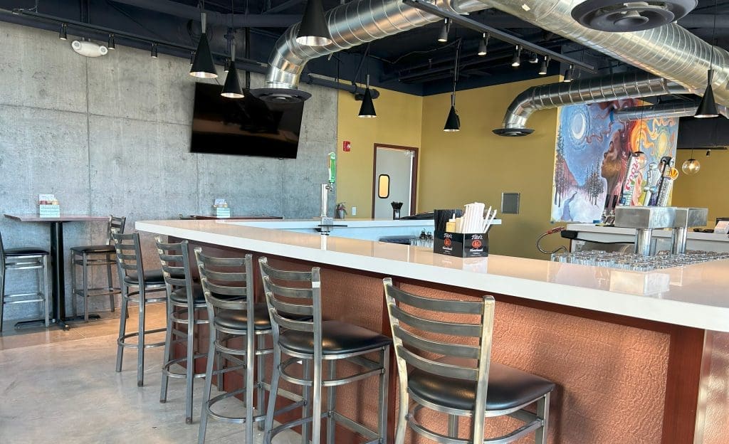 Modern bar interior with metal stools, a white countertop, exposed ductwork, concrete walls, a TV, and colorful wall art. The bar is empty and well-lit by natural light.