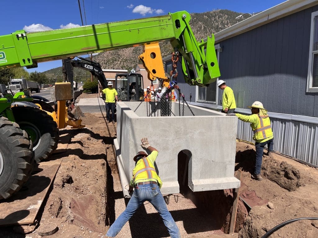 Construction workers guide a large concrete structure being lowered by a green crane into a trench next to a building on a sunny day.