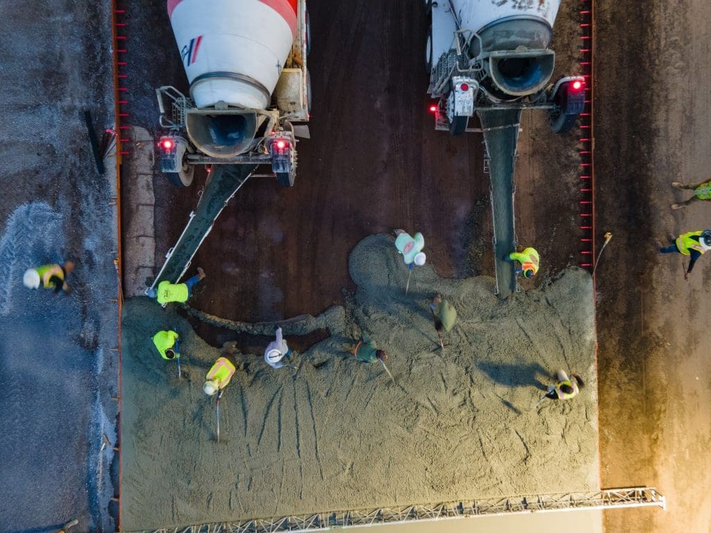 Aerial view of construction workers spreading fresh concrete from two cement mixer trucks onto a roadway.