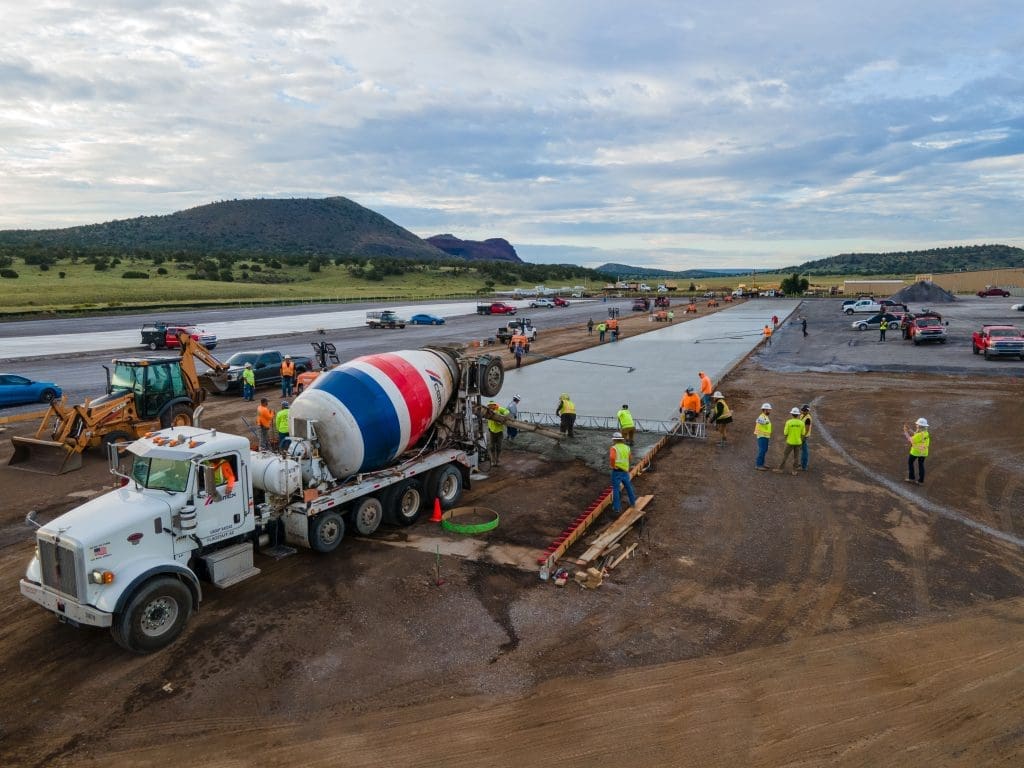 Construction workers pour and smooth concrete on a large outdoor worksite with heavy machinery, vehicles, and hills in the background.