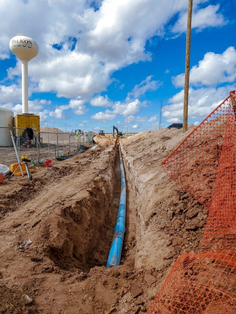 A blue utility pipe is laid in a trench at a construction site, with dirt piles, orange safety fencing, and a water tower in the background under a partly cloudy sky.