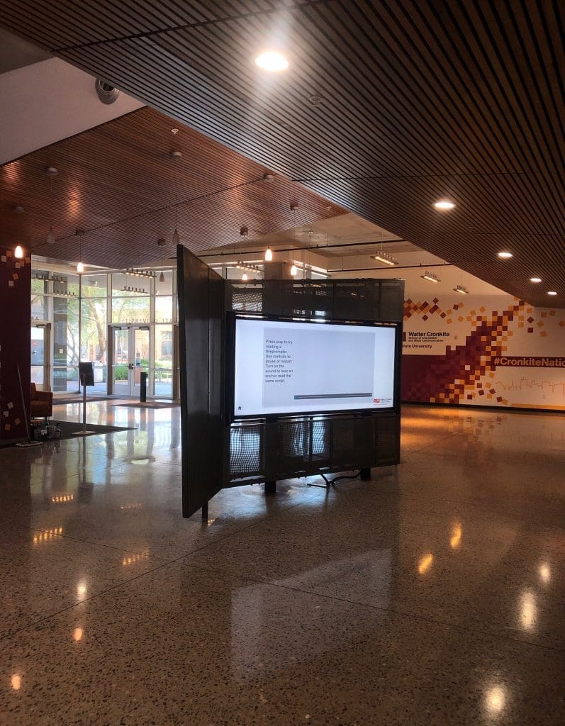 A large digital display stands in the center of a spacious lobby with terrazzo flooring, wood-paneled ceiling, and mosaic wall art.