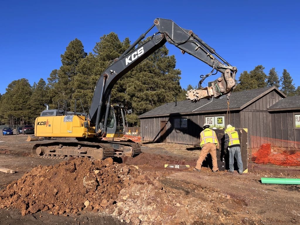 An excavator with a KCS logo is parked on a construction site while two workers in safety vests stand next to a small building.