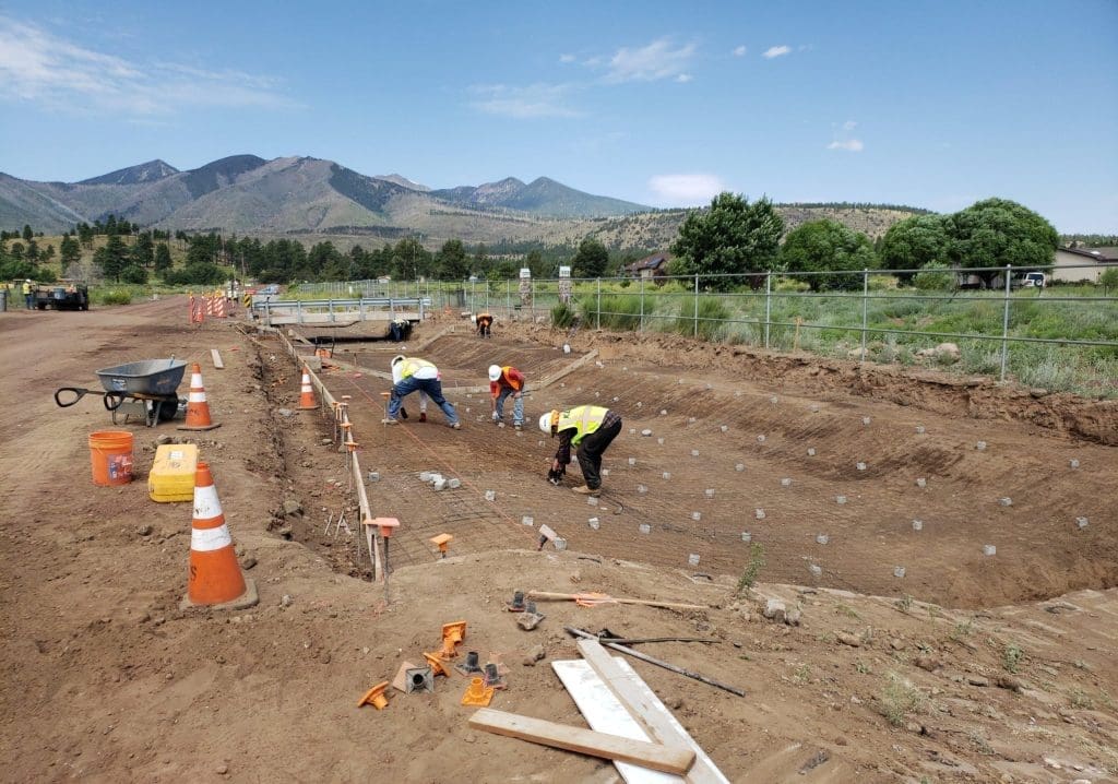 Several construction workers install rebar in a large, excavated dirt pit at an outdoor construction site with mountains in the background.