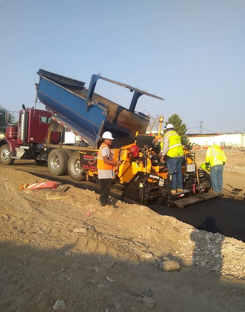 Construction workers operate paving machinery as asphalt is poured from a dump truck onto a road under construction at a worksite.