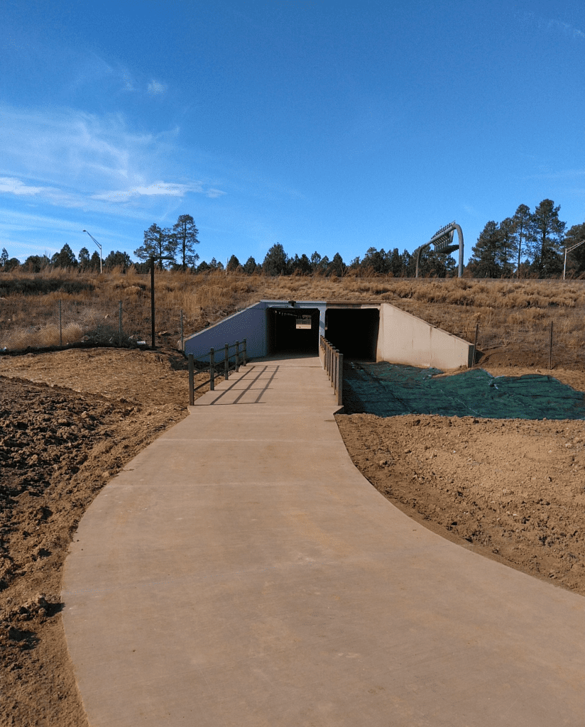 A paved pathway leads to an underpass tunnel beneath a road, surrounded by dry grass, trees, and a clear blue sky.