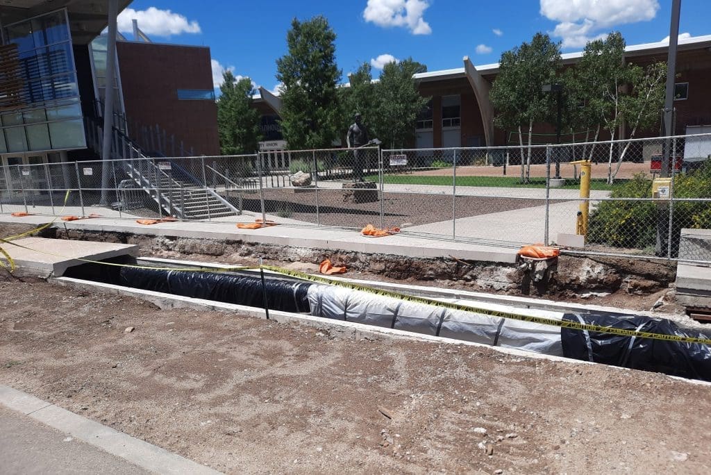 A long trench is dug along a sidewalk on a campus, bordered by construction fencing and caution tape, with buildings and trees in the background.