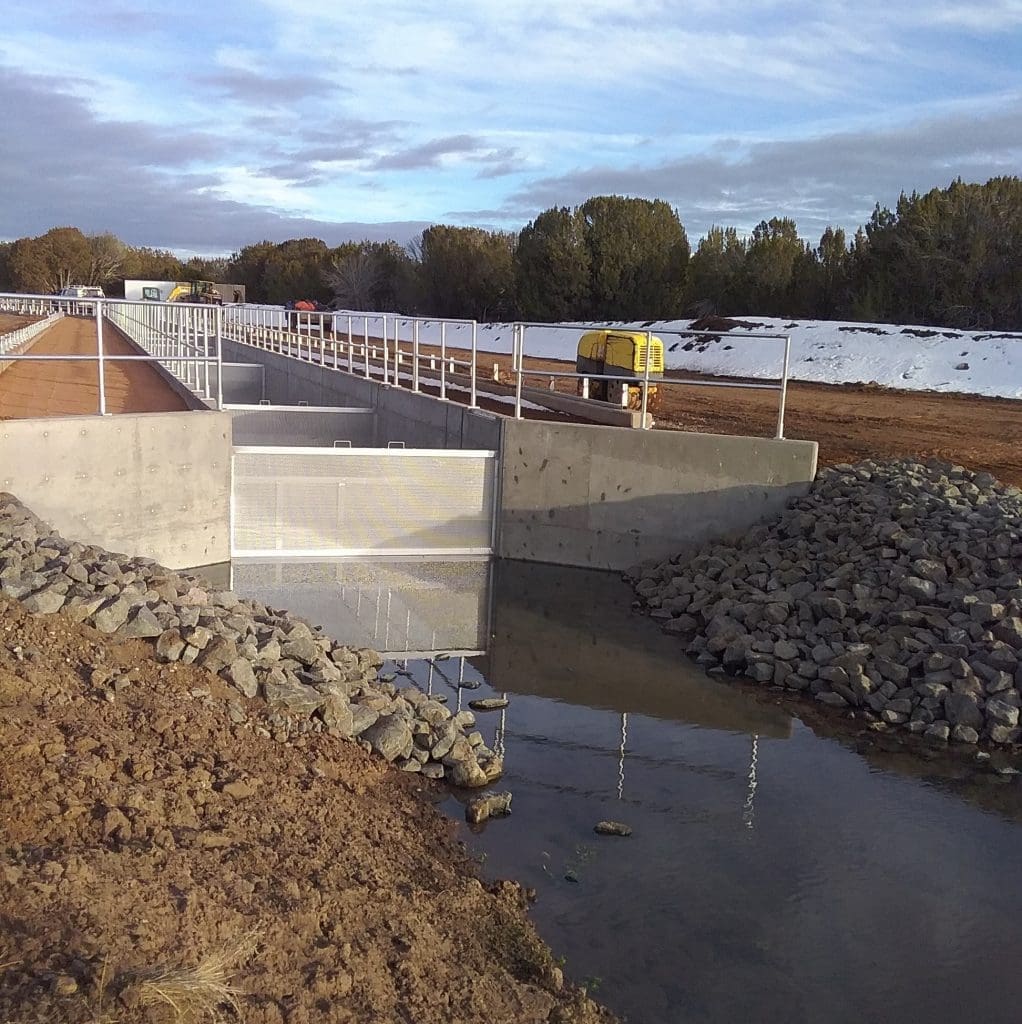 A concrete irrigation structure with metal railings channels water, with construction equipment and rocks nearby; snow and trees are visible in the background.