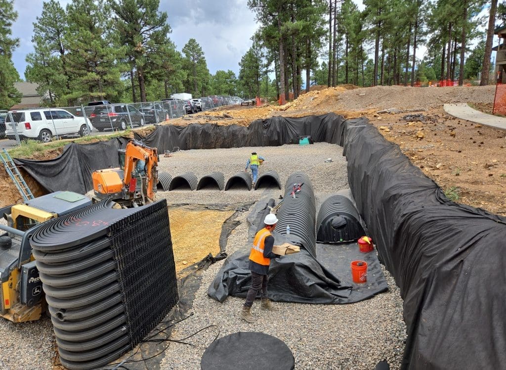 Construction workers install large black plastic drainage pipes and structures in a deep excavation site, surrounded by gravel and lined with black fabric, near a forested area.