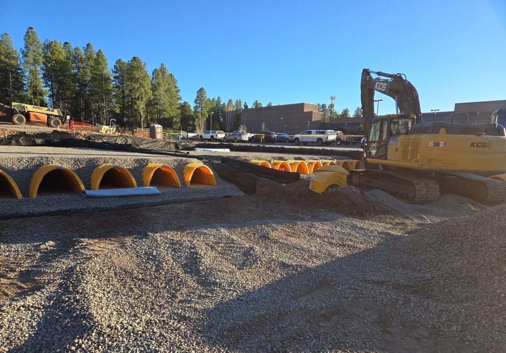 Construction site with a yellow excavator and multiple large yellow drainage pipes partially buried in gravel, with vehicles and trees in the background.