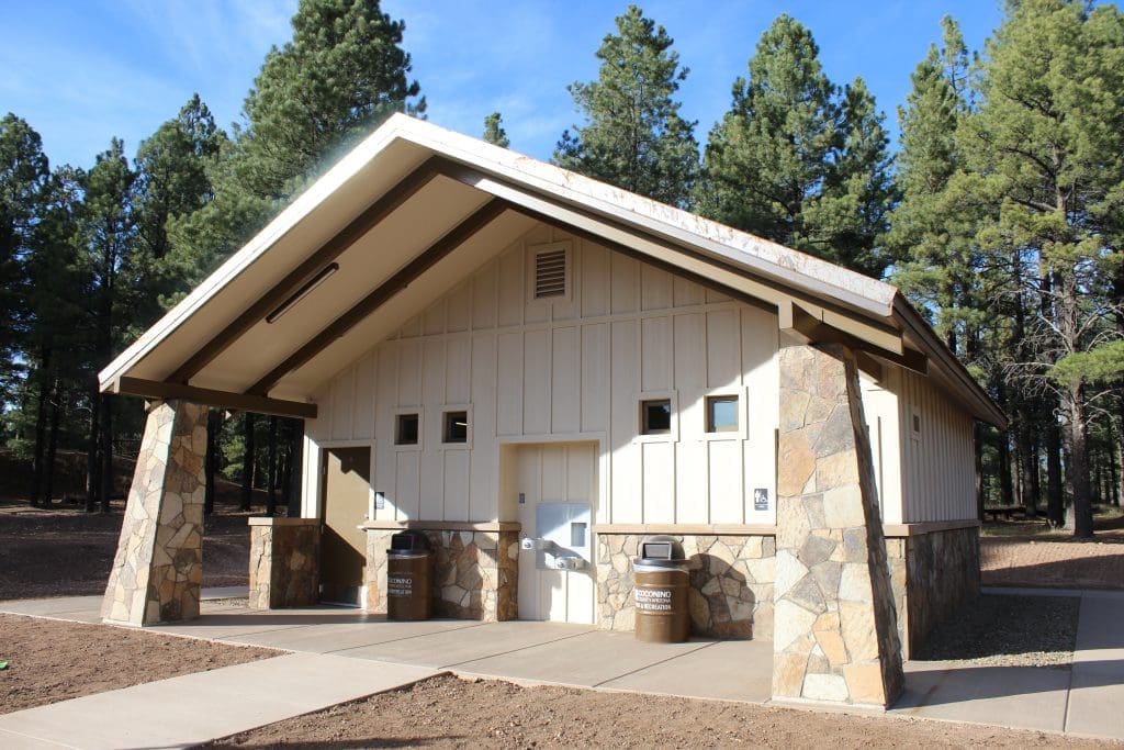 A public restroom building with a stone and wood exterior, located in a forested area with tall pine trees in the background.
