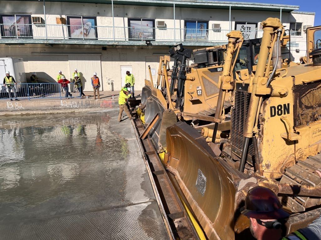 Construction workers operate bulldozers and smooth wet concrete at a building site with multiple workers and equipment visible.