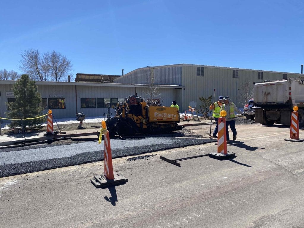 Construction workers operate machinery and lay asphalt on a road in front of a commercial building, with traffic barriers set up around the work area.