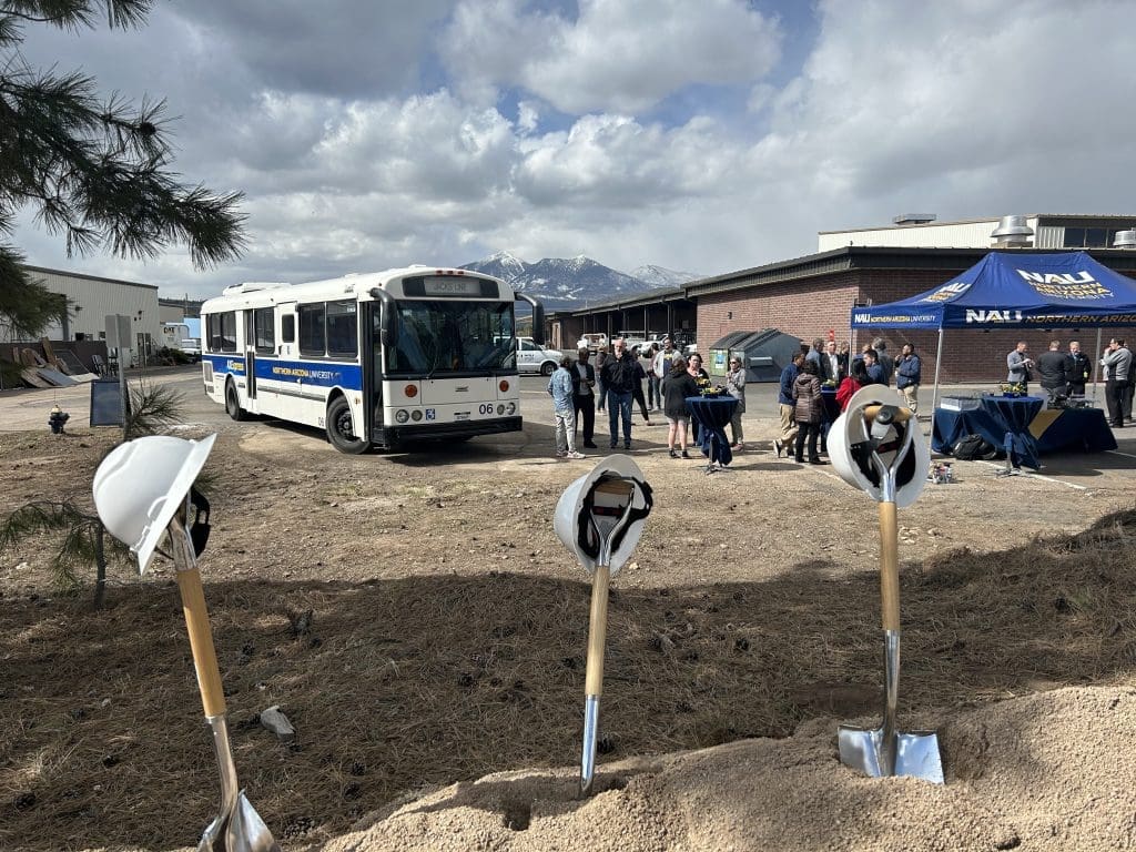 A group of people gather near a parked bus, shovels, and hard hats at the Kinney’s Mountain Line CDL Training Course Project Groundbreaking Ceremony outside a building with mountains in the background.