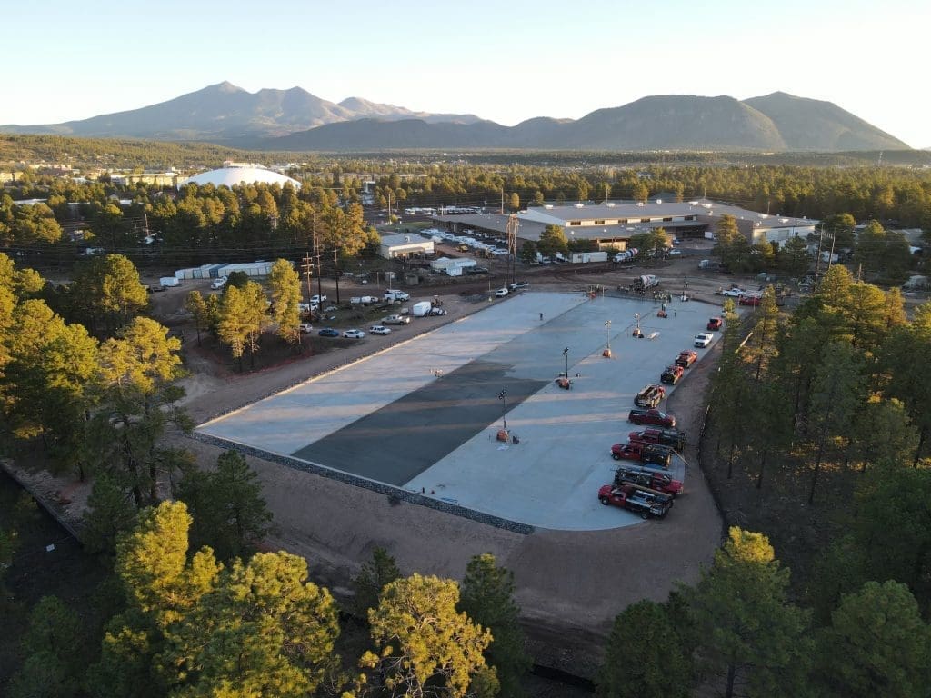 Aerial view of a large concrete slab being poured at a construction site surrounded by trees, with vehicles and workers present and mountains visible in the background.