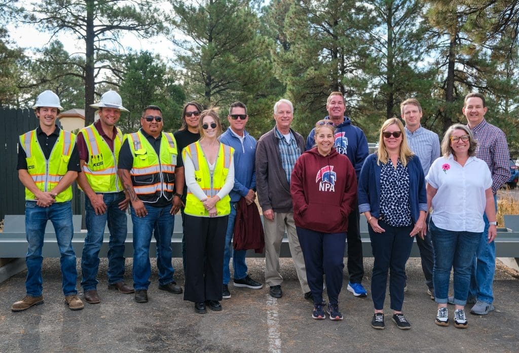 A group of thirteen people, including construction workers in safety vests and others in casual clothing, stand outdoors on pavement with trees behind them as the Northland Preparatory Academy STEAM Building reaches a key construction milestone.