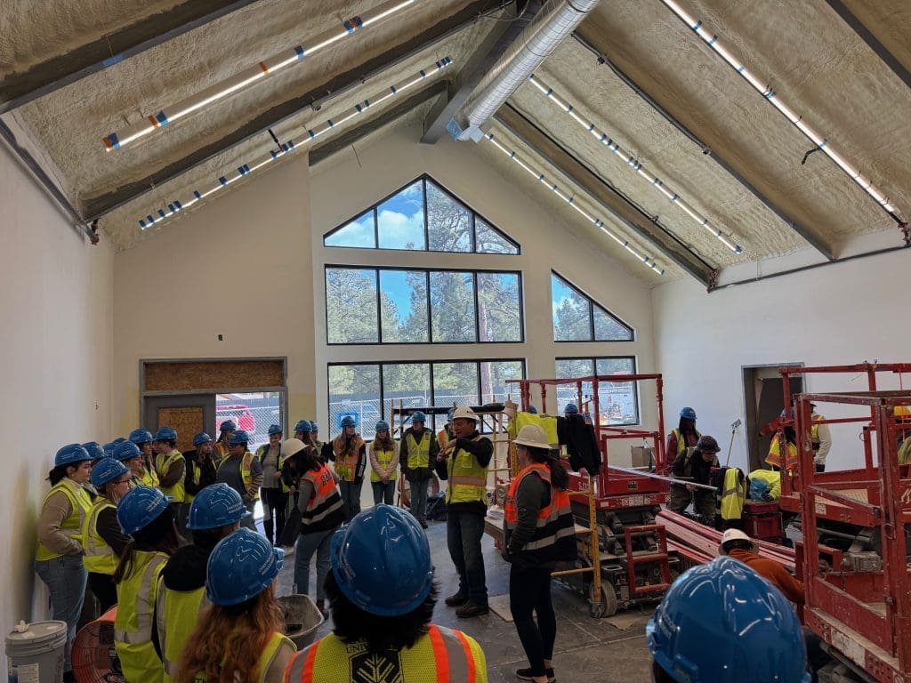 Inspiring the Next Generation: Interior Design Students visit a Kinney job site, where they gather in construction vests and hard hats inside a partially finished building with exposed ceiling insulation and large windows.