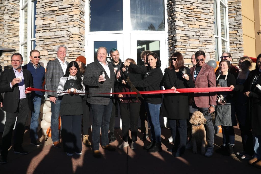 A group of people stand in front of Noble Ground Coffee during a ribbon-cutting ceremony; a woman cuts the red ribbon while others hold drinks and smile, celebrating Flagstaff's newest coffee shop with a cause.