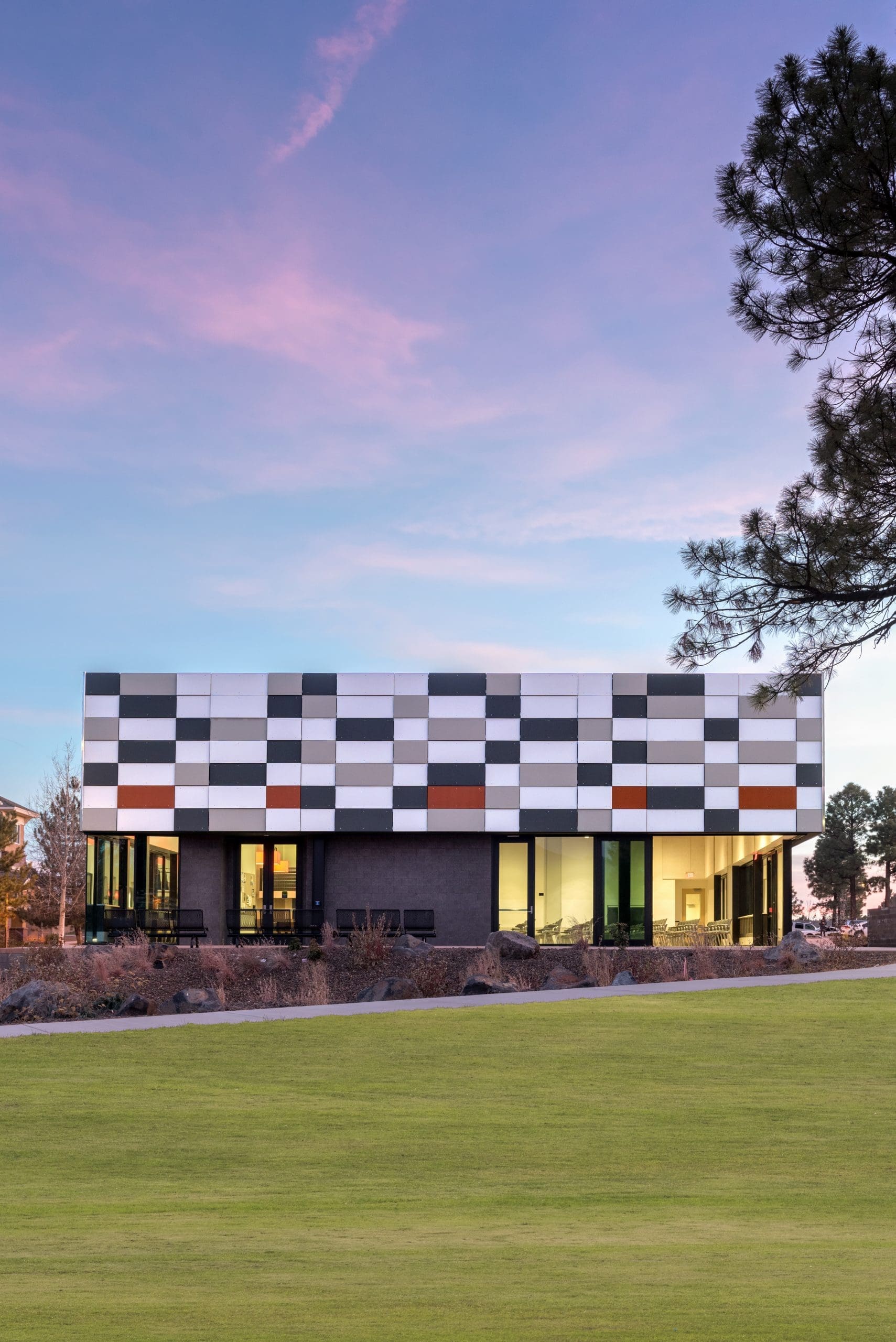 Modern building with a checkered pattern facade, large windows, and a green lawn in front, set against a sunset sky and framed by pine trees.