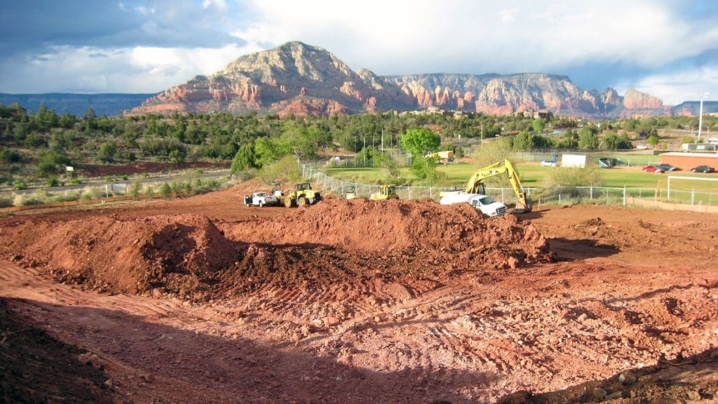 Construction vehicles and equipment at a red dirt excavation site, where the Sedona-Oak Creek USD PV System Receives Award of Excellence, with mountains and green trees in the background under a partly cloudy sky.