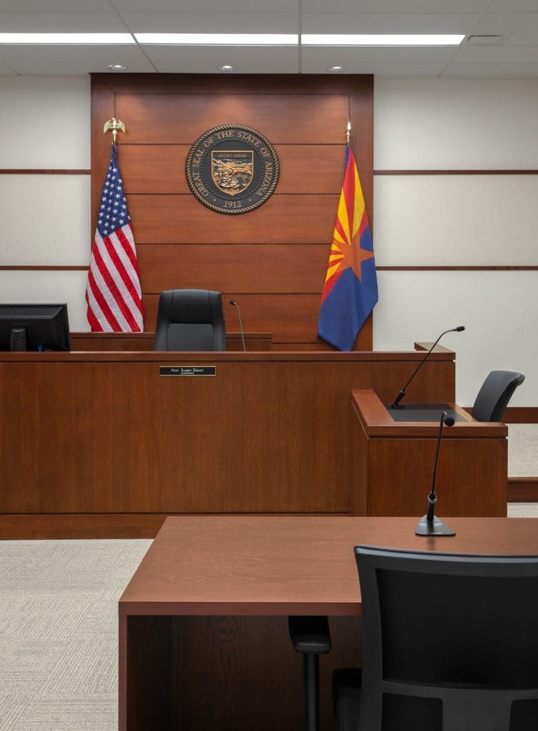View of an empty City of Flagstaff Municipal Court Facility courtroom with judge’s bench, witness stand, two flags, and the seal of the State of Arizona on the wall.