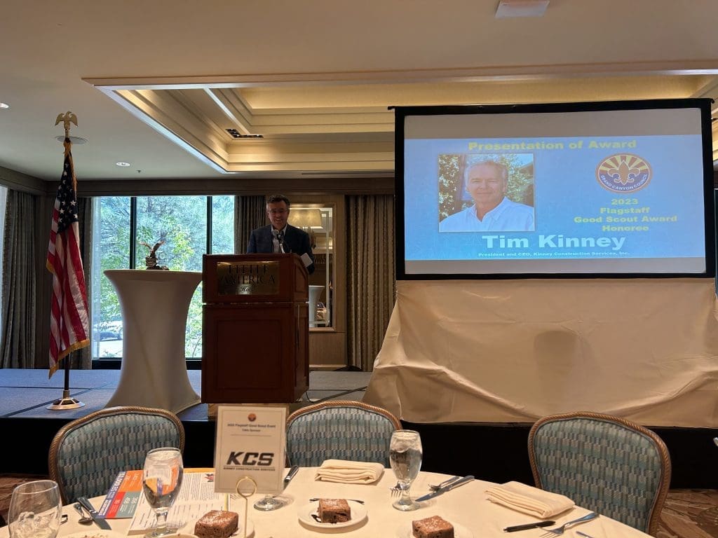 A man stands at a podium giving a presentation in a banquet room, with a projection behind him announcing Tim Kinney as the 2023 Good Scouts' Award Recipient.