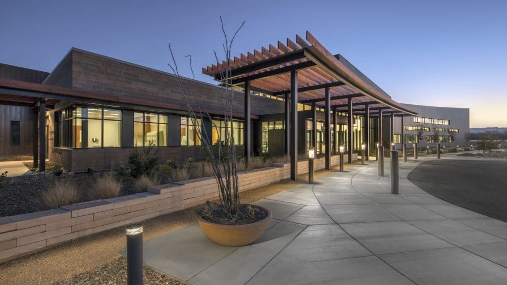 Modern building with large windows and a covered walkway at dusk, surrounded by landscaped planters and exterior lighting, as seen at Verde Valley Medical Center – Camp Verde Campus, recipient of Arizona Masonry Guild’s Excellence in Masonry Architectural Award.