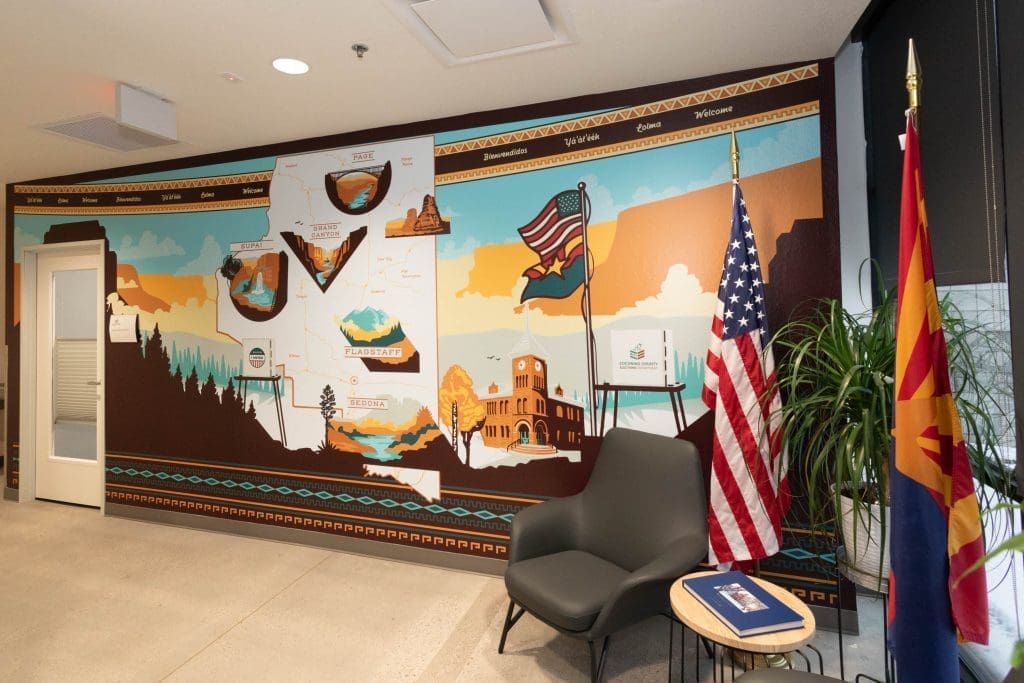A mural depicting Arizona landmarks and a map on a wall, next to a gray chair, a small table with a booklet, two flags, and potted plants in an office space.