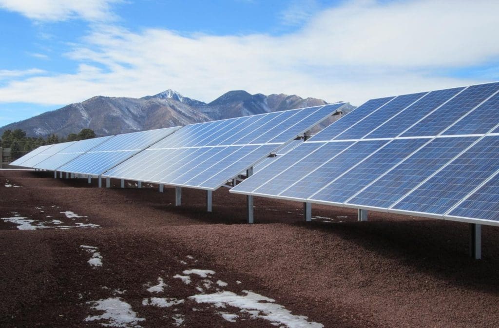 Rows of solar panels from the APS Doney Park PV System are installed on the ground, with mountains and a partly cloudy sky in the background.