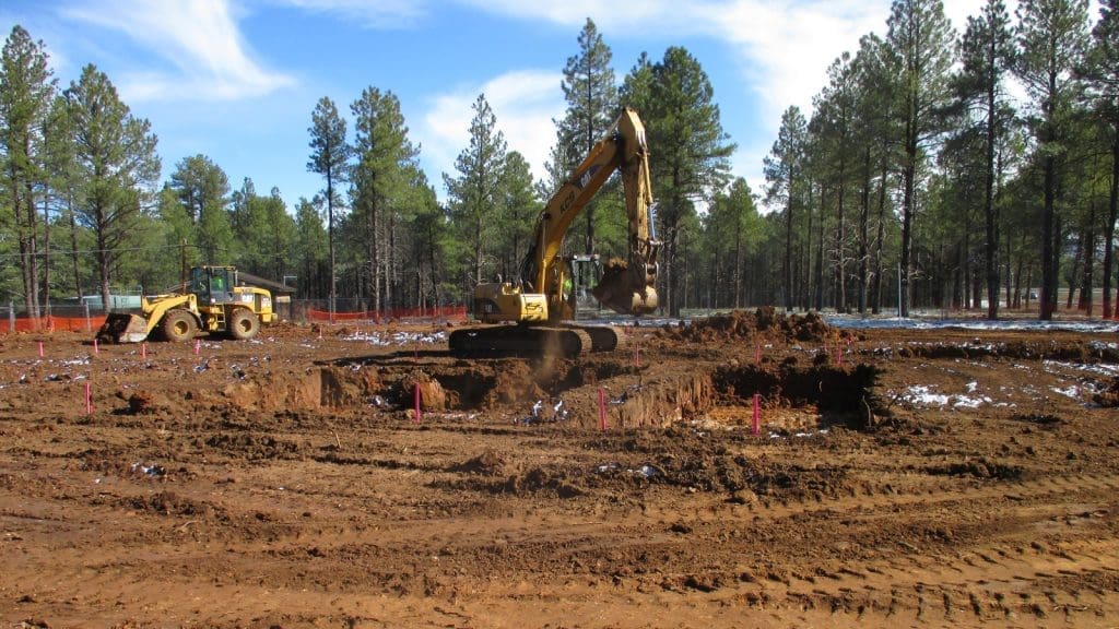 An excavator digs a large hole at a construction site in a forested area, with a bulldozer and orange safety fencing in the background.