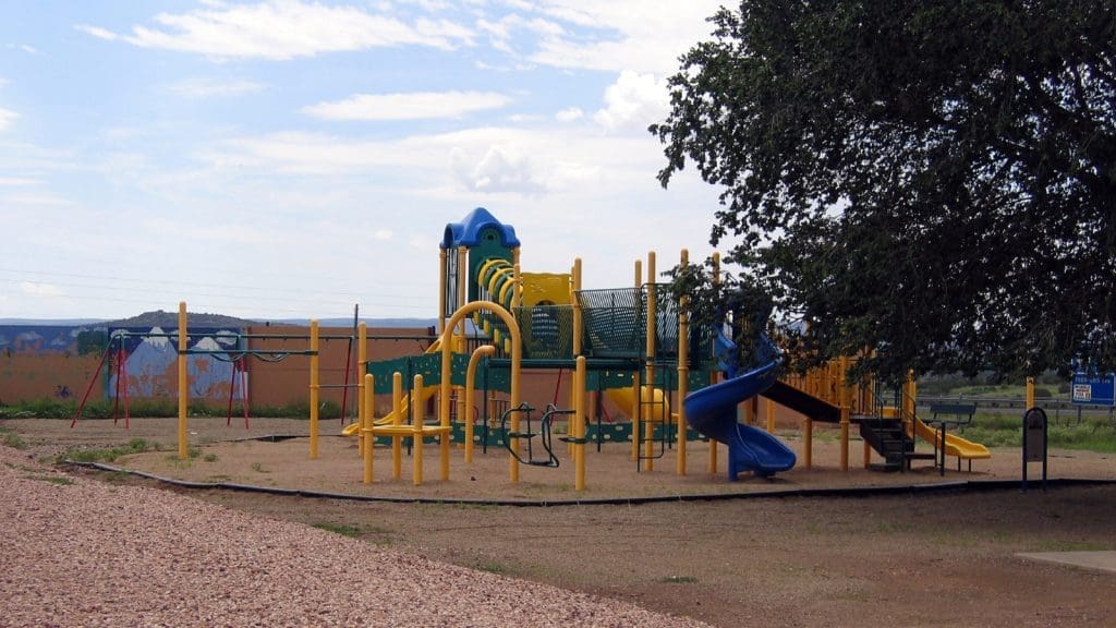A playground with yellow and green climbing structures, slides, and bridges sits on a sandy surface beside a large tree under a partly cloudy sky.