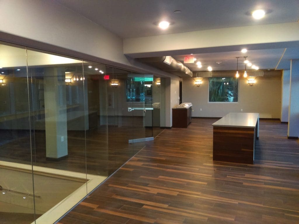Modern office space with glass walls, dark wood flooring, overhead lights, and large desk, featuring an open layout with windows and visible outside greenery.