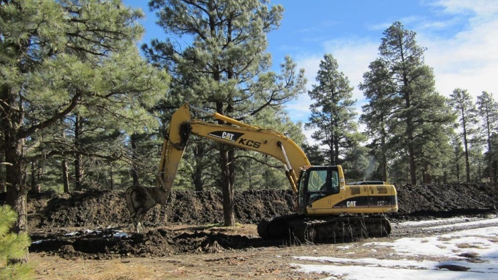 A yellow CAT excavator is digging soil in a forested area with pine trees and patches of snow on the ground.