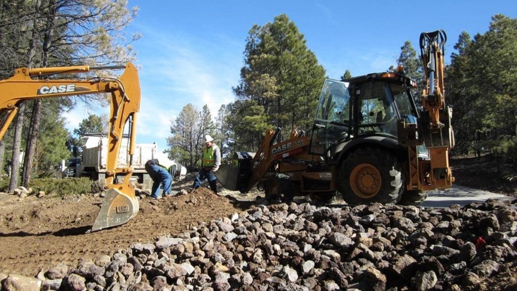 Construction workers operating heavy machinery and working on a dirt road near a forested area with rocks in the foreground.