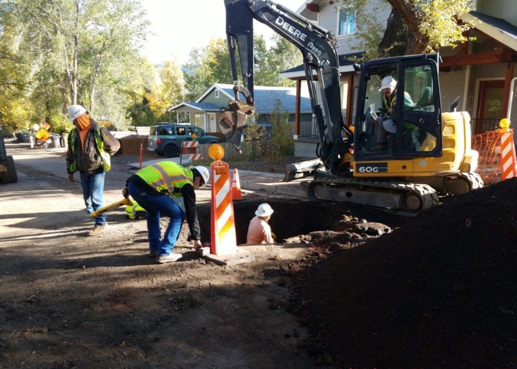 Construction workers operate machinery and work in a large hole on a residential street, surrounded by safety barriers and traffic cones.