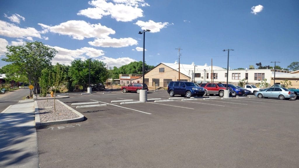 A mostly empty parking lot with a few cars parked, surrounded by trees and buildings under a blue sky with scattered clouds.