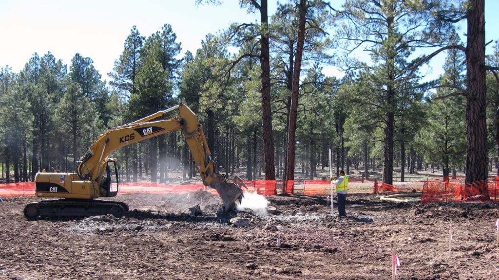 An excavator digs in a forested area while a construction worker in a safety vest observes nearby; orange safety fencing surrounds the site.